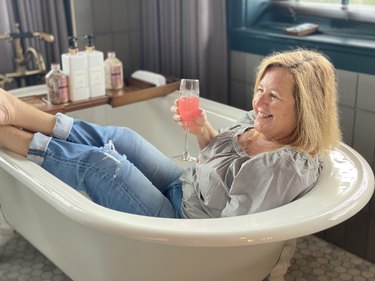 A woman sits in a bathtub holding a pink beverage in a champagne flute