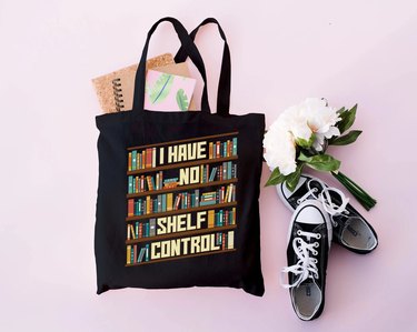 A black tote bag featuring a tilted bookshelf image with the words "I have no shelf control." The bag is sprawled out on a pink background next to a pair of black Converse sneakers, a pile of journals and a white floral bouquet.