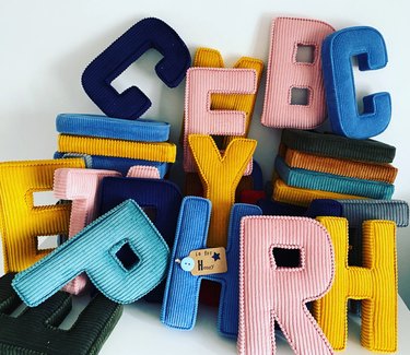 A pile of various corduroy letters in pink, mustard yellow, dark blue, teal, brown and forest green, including a blue letter "H" with a brown tag reading "H is for Henry"