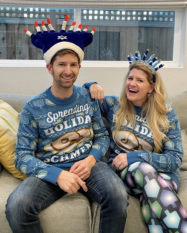 A man and a woman sit side by side on a couch wearing menorah hats and matching blue sweaters that read "Spreading Holiday Schmear."