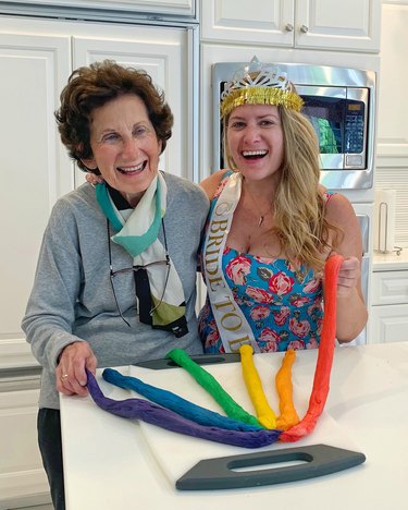 Two women, one in her 90s and one younger, smile while holding strands of rainbow-colored dough