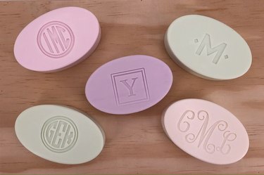 Five oval-shaped bars of soap engraved with various letters. The soap bars are pastel pink, purple and green.