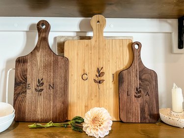 A set of three bamboo wood cheese boards with letters and leaves engraved in the center. A light pink flower sits in front of the cheese boards.