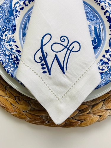 A white napkin embroidered with a navy blue cursive letter "W." The. napkin sits on a blue and white plate and woven wood placemat.