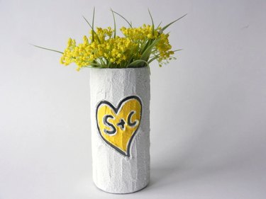 A white cylindrical vase with a yellow heart and "S+C" carved in the center. There are dainty yellow flowers in the vase.