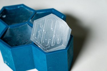 A deep blue silicone mold shaped like four adjoined hexagons. One hexagon has a monogrammed hexagonal ice cube emerging from the top.
