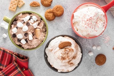 Three dessert holiday dips in colorful mugs topped with whipped cream, marshmallows, crushed-up peppermints and chocolate chips.