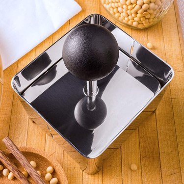 Stainless steel Raw Rutes tofu press, shown in overhead view on a butcher block countertop with soybeans visible above and below it