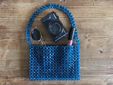 Blue beaded bag on a wood backdrop with a camera, lipstick and sunglasses coming out of the bag