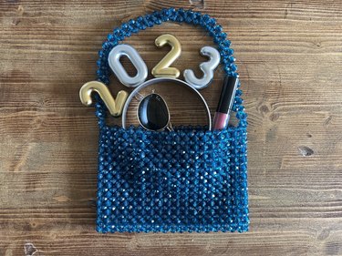 A blue beaded bag against a wood background with sunglasses, lip gloss and a 2023 party hat coming out of the bag