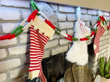 A closeup of three Christmas stockings hanging above a gray brick fireplace with a red, green, and white chain garland.