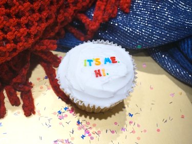 A white cupcake with the phrase "It's me. Hi." spelled out in multicolored edible letters.