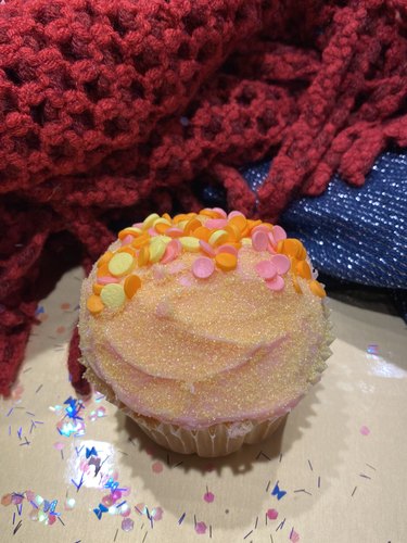 A cupcake with pink frosting; yellow edible glitter; and pink, orange and yellow circular sprinkles.