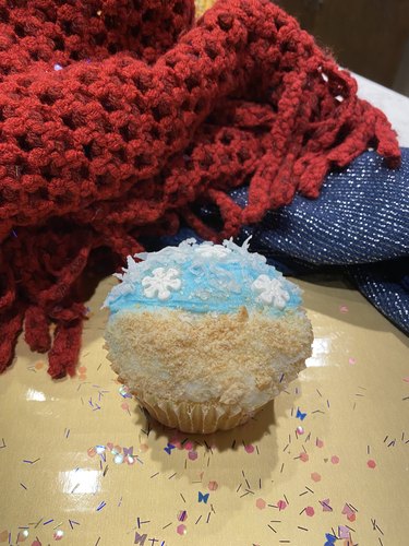 A cupcake with light blue frosting, graham cracker crumbs made to look like sand and coconut flakes with snowflake candy atop the blue frosting.