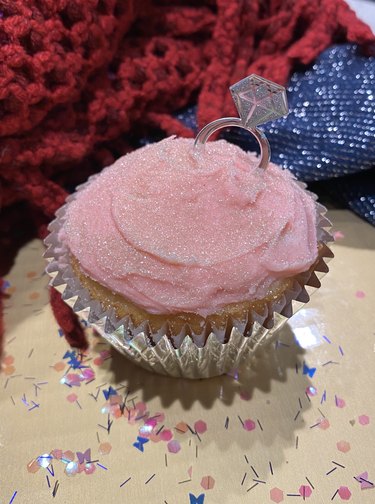 A cupcake with pink frosting, edible silver glitter and a paper diamond ring stuck into the top.