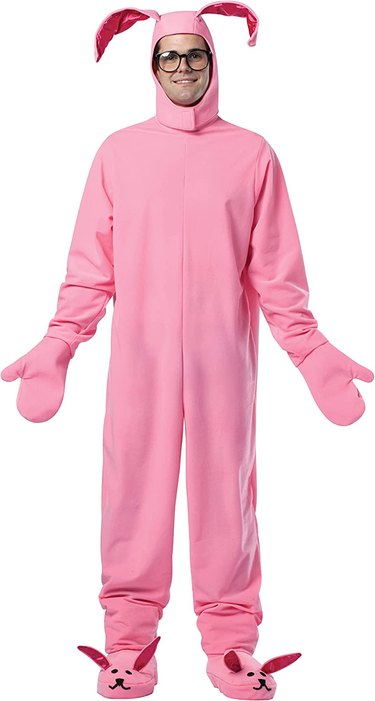 A man with glasses wears a bright pink onesie with bunny ears, mitten hands and bunny feet.
