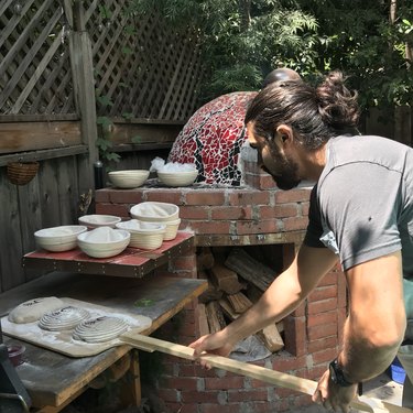 Man holding three loaves of bread on a wood platter next to a red-tiled bread oven