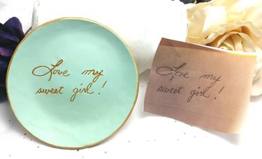 Light blue clay dish engraved in gold with "Love my sweet girl!" next to a piece of paper with the same words written in pen