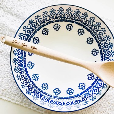 White and blue patterned bowl under a light wooden serving spoon. The spoon's handle is engraved with "Love you, Mom"