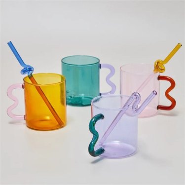 Handmade two-tone clear glass mugs with wavy handles that resemble the shape of an ear and curved, whimsical glass straws to match.