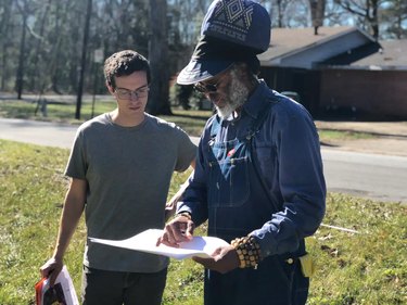 Black man with large blue hat and young white man in gray T-shirt look over plans for a garden on paper