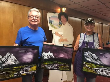 Standing on either side of a cardboard cutout of artist Bob Ross, a man in blue shirt and a woman wearing gray tank top and purple apron hold three paintings of mountains.
