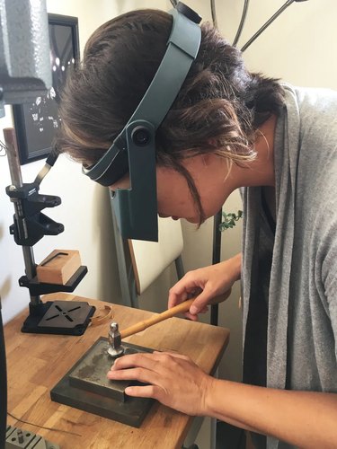 Person wearing gray cardigan and protective goggles works with sterling silver hammer