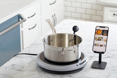 Hestan Cue induction cooker, pictured on a marble counter with a pot, temperature probe and smartphone displaying the Hestan app