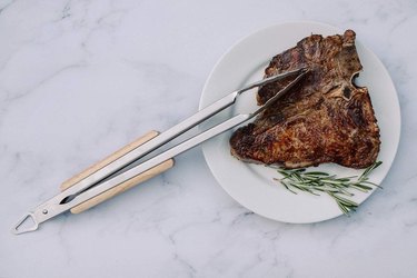 Grilling tongs resting on a grilled steak, on a white plate, on a marble counter