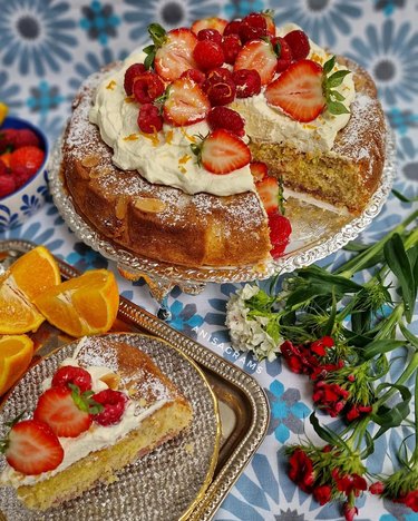 Cake topped with white frosting, fresh strawberries and fresh raspberries, next to a plate of cut-up oranges