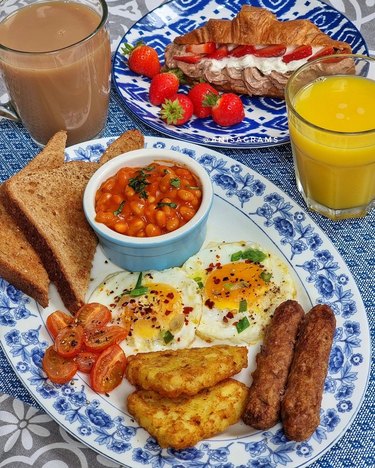 Full English breakfast plate with sausage, eggs, toast, tomatoes, potatoes, orange juice, a stuffed croissant and masala beans