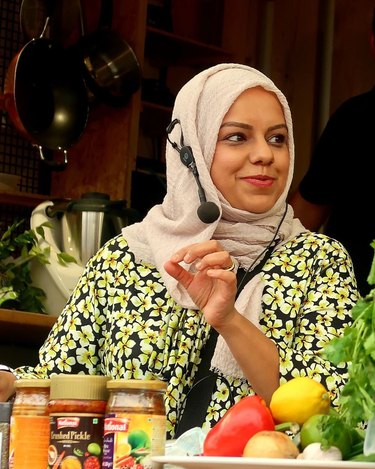 Woman in floral dress and tan hijab speaks into a microphone