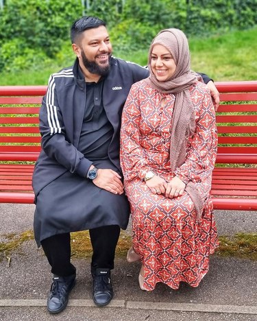 Woman in red and blue patterned dress and light brown headscarf sits next to man in striped black and white jacket with black tunic on a red bench