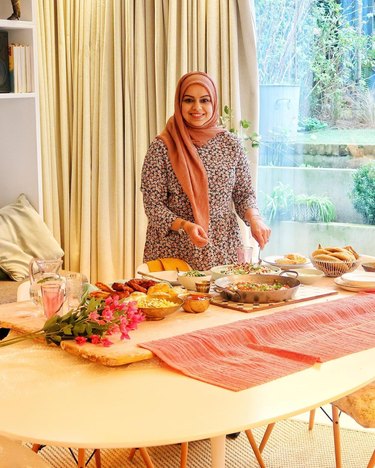 Woman in patterned dress and pink headscarf stands over a table with various plates of food