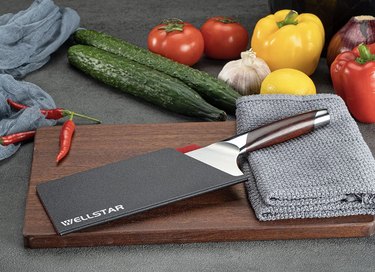 Plastic knife cover by Wellstar for a cleaver knife on a walnut cutting board surrounded by vegetables and a kitchen towel.