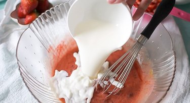 Pouring milk into a large glass mixing bowl with pink donut batter