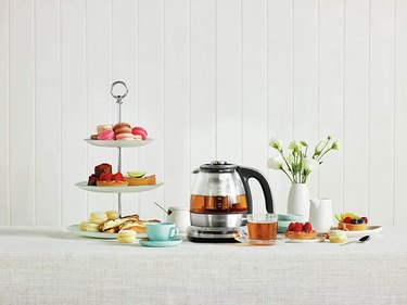 The Breville Smart Tea Infuser sitting on a table with black tea in it, surrounded by teacups, small desserts, and flowers.