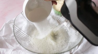 Glass mixing bowl with foamy egg whites. Baker is slowly pouring in granulated sugar while mixing.