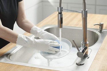 A pair of hands at a sink wearing white cleaning gloves, scrubbing a clear dish with a blue sponge.