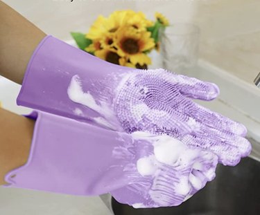 A pair of hands wearing light purple silicon gloves, covered in bubbles and washing up at a sink. there is a vase full of sunflowers in the background.