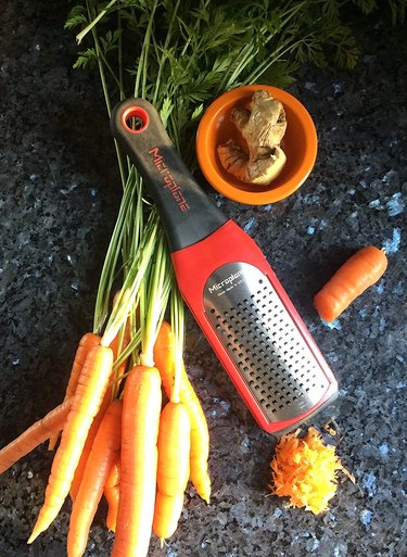 A Microplane Artisan Series Coarse Cheese Grater on a countertop surrounded by carrots and ginger
