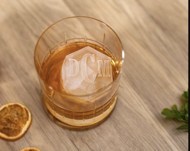 Monogrammed, hexagon-shaped ice cube in a whiskey tumbler on a wood countertop.