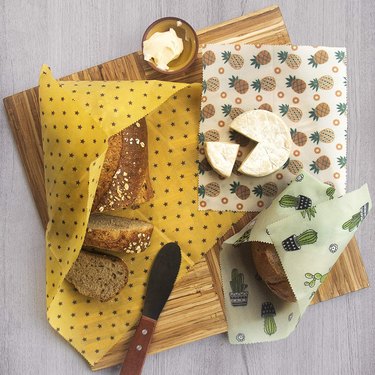 Beeswax wraps in star, pineapple, and cactus patterns on a wood cutting board holding cheese and bread.