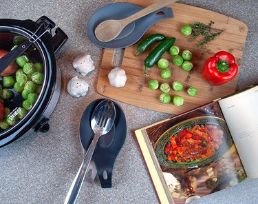 Two silicone spoon rests—one navy blue and one blue/gray—on a countertop next to a cutting board, Crockpot, and cookbook.