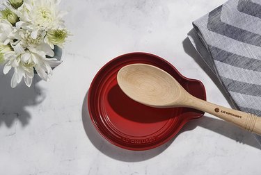 Le Creuset stoneware spoon rest in red on a marble countertop next to flowers and a striped kitchen towel.