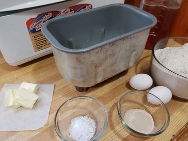 Bread pan from a Zojirushi bread machine, shown on a counter with ingredients (honey, wheat flour, eggs, yeast, salt and butter) arranged around it in a half-circle.