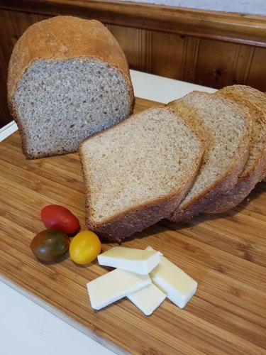 A loaf of fresh bread, half-sliced, shown on a bamboo cutting board with pieces of aged cheddar and a few colorful cherry tomatoes