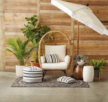 Rattan, natural-colored egg chair with off-white back, side, and seat cushions surrounded by decor like a fringed umbrella, pouf, area rug, and potted plants.