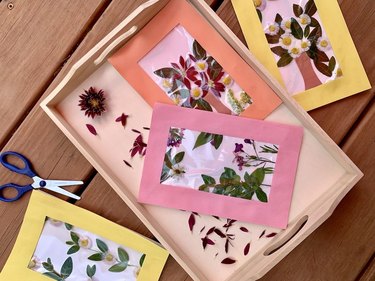 Completed Mother's Day cards with frames in yellow, pink, and orange sit atop a wooden table and tray and feature pressed flowers