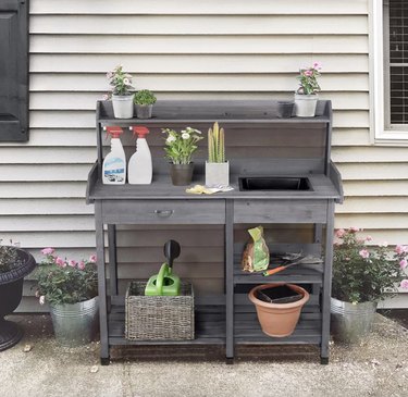 Loon Peak Fir Potting Bench in Gray Pushed Up Against the Side of a House With Supplies on the Shelves